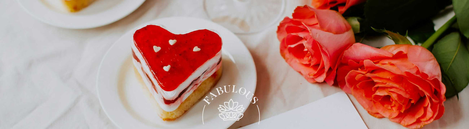 A heart-shaped strawberry cake with white heart decorations on a plate next to vibrant pink roses, creating a romantic and delightful setting - Fabulous Flowers and Gifts.