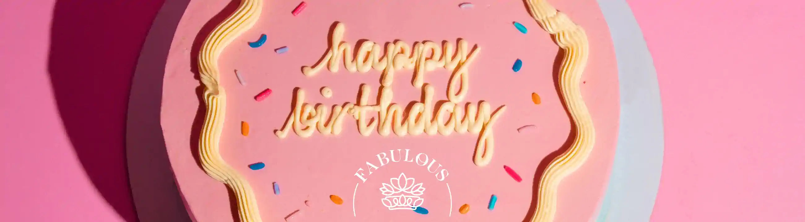 A pink birthday cake with the words "happy birthday" written in white icing, decorated with colorful sprinkles. Fabulous Flowers and Gifts