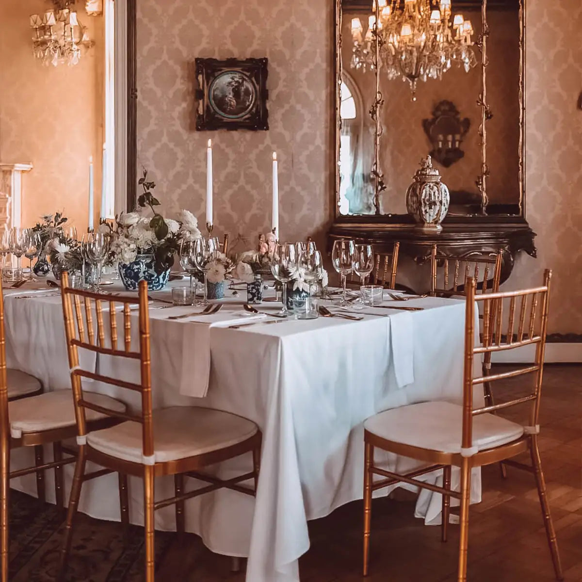 Luxurious event dining room with a round table adorned with white linens, gold chairs, tall candles, and elegant floral arrangements, set against a backdrop of ornate decor and chandeliers. Fabulous Flowers and Gifts.