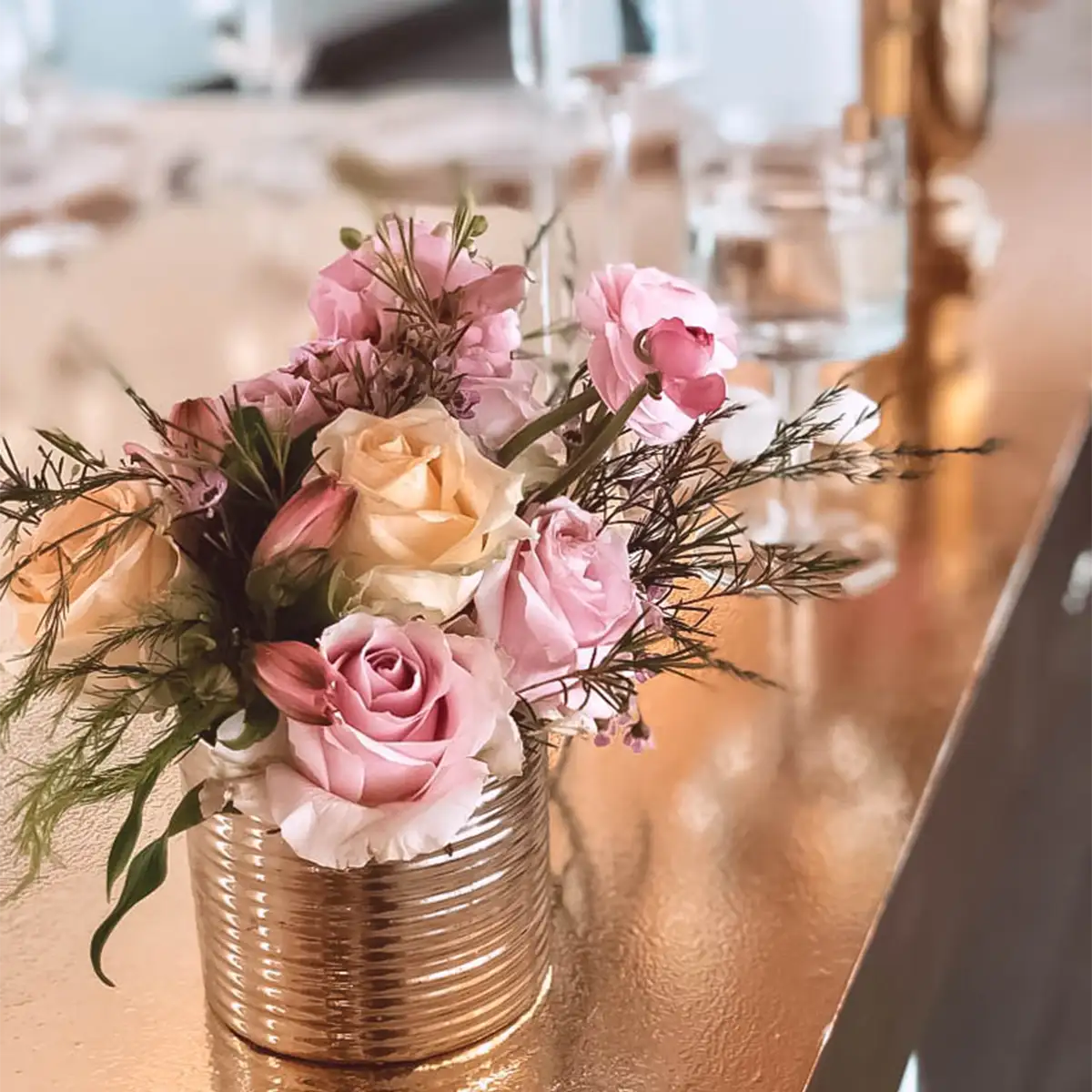 Elegant small floral arrangement with pink and cream roses in a gold vase, set on a reflective gold table with glassware in the background. Perfect for event decor. Fabulous Flowers and Gifts.