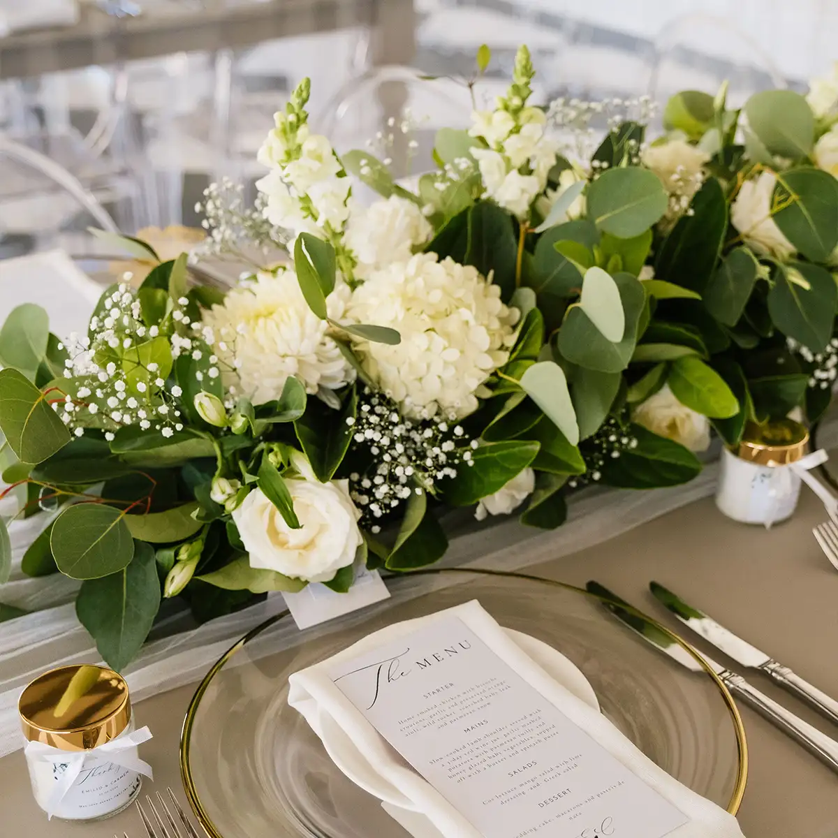 Elegant event table setting featuring a lush green and white floral centrepiece, clear glass plates with gold accents, and a beautifully printed menu. Perfect for sophisticated events. Fabulous Flowers and Gifts.