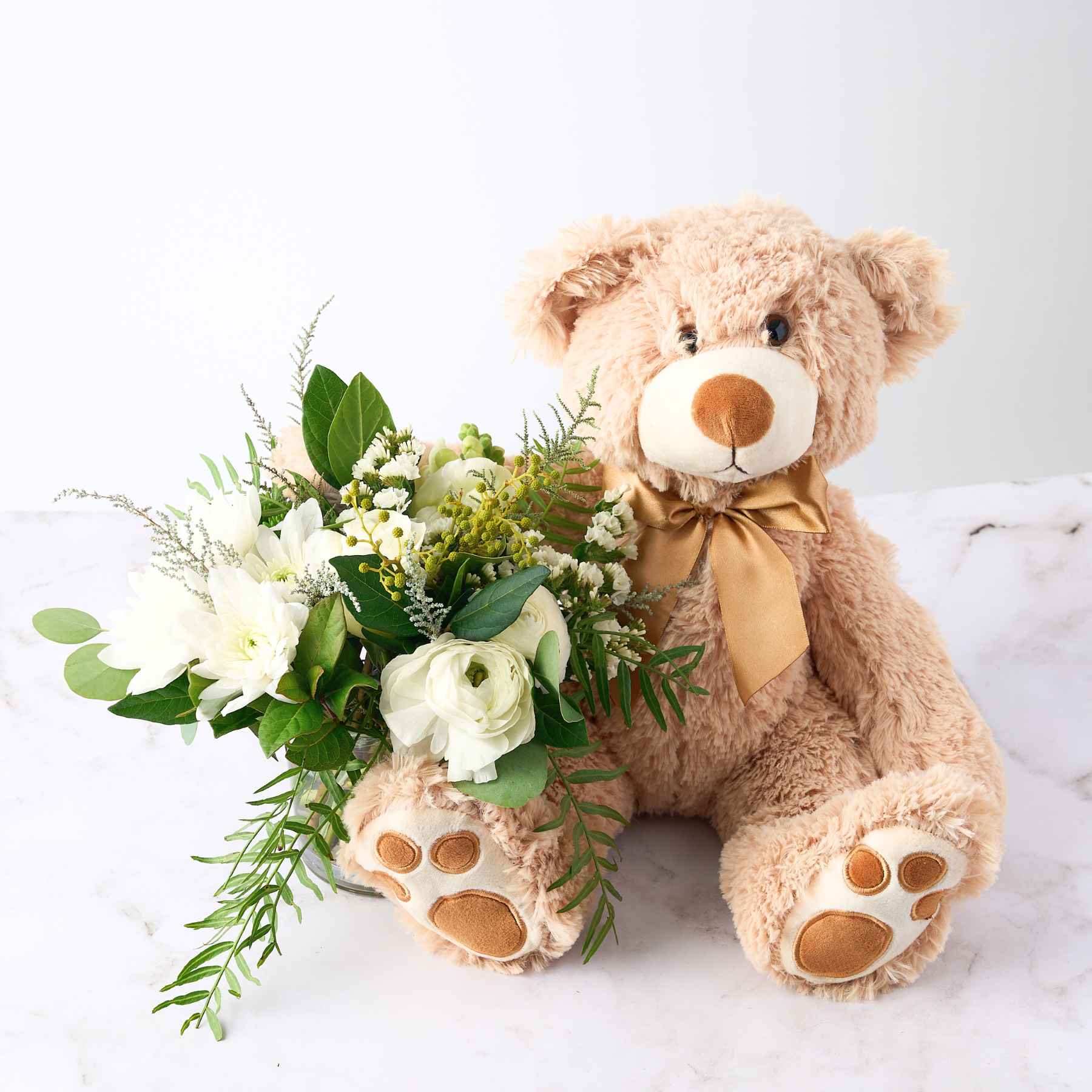 A plush teddy bear with a golden ribbon, sitting beside a bouquet of white flowers and greenery, representing the charming 'A Baby Has Arrived' gift arrangement.