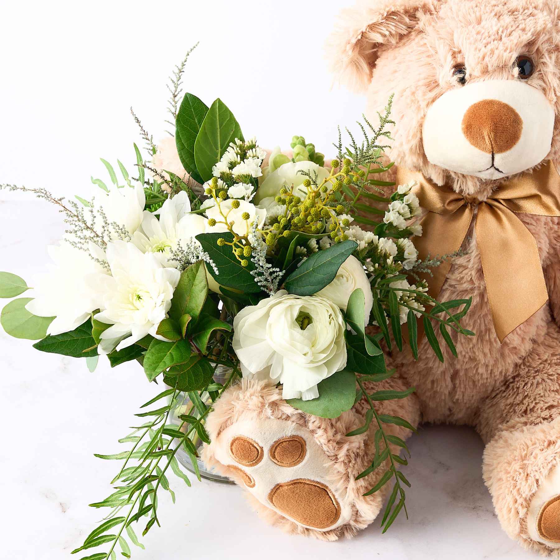 Close-up of a teddy bear with a bouquet of white flowers and green foliage, capturing the joyful essence of the 'A Baby Has Arrived' gift.