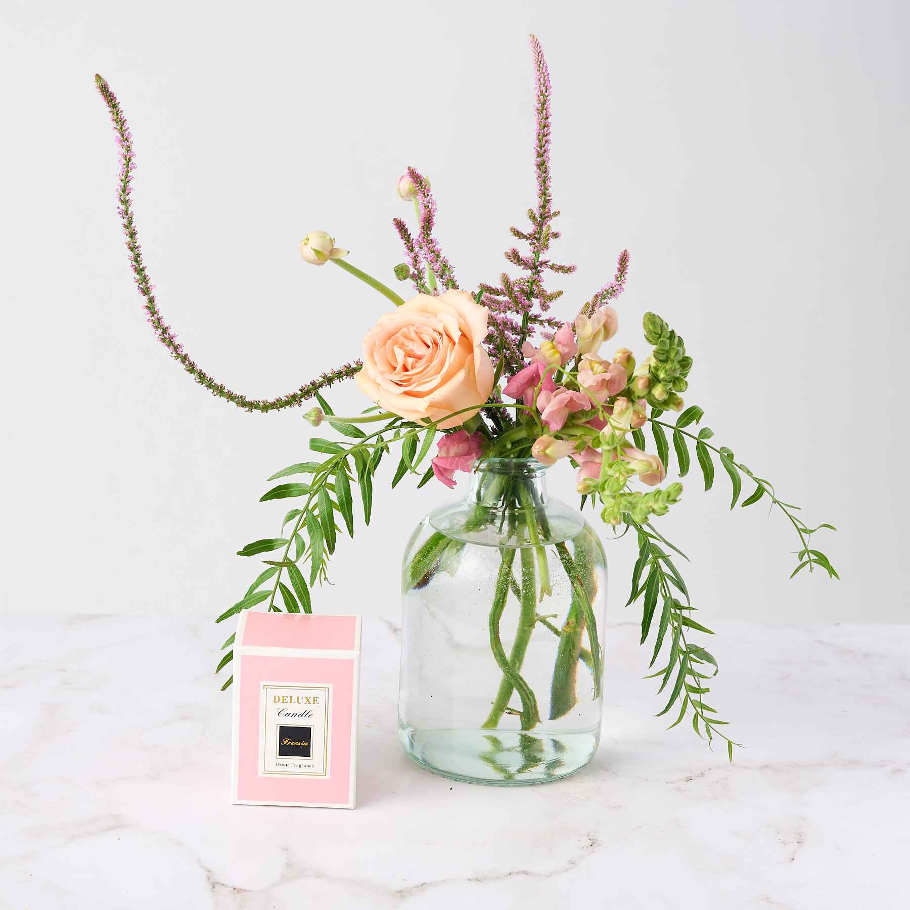 A delicate flower arrangement in a glass vase featuring soft peach roses and pink blooms, paired with a pink deluxe candle box, part of the 'A Moment of Calm Pamper Gift Set'