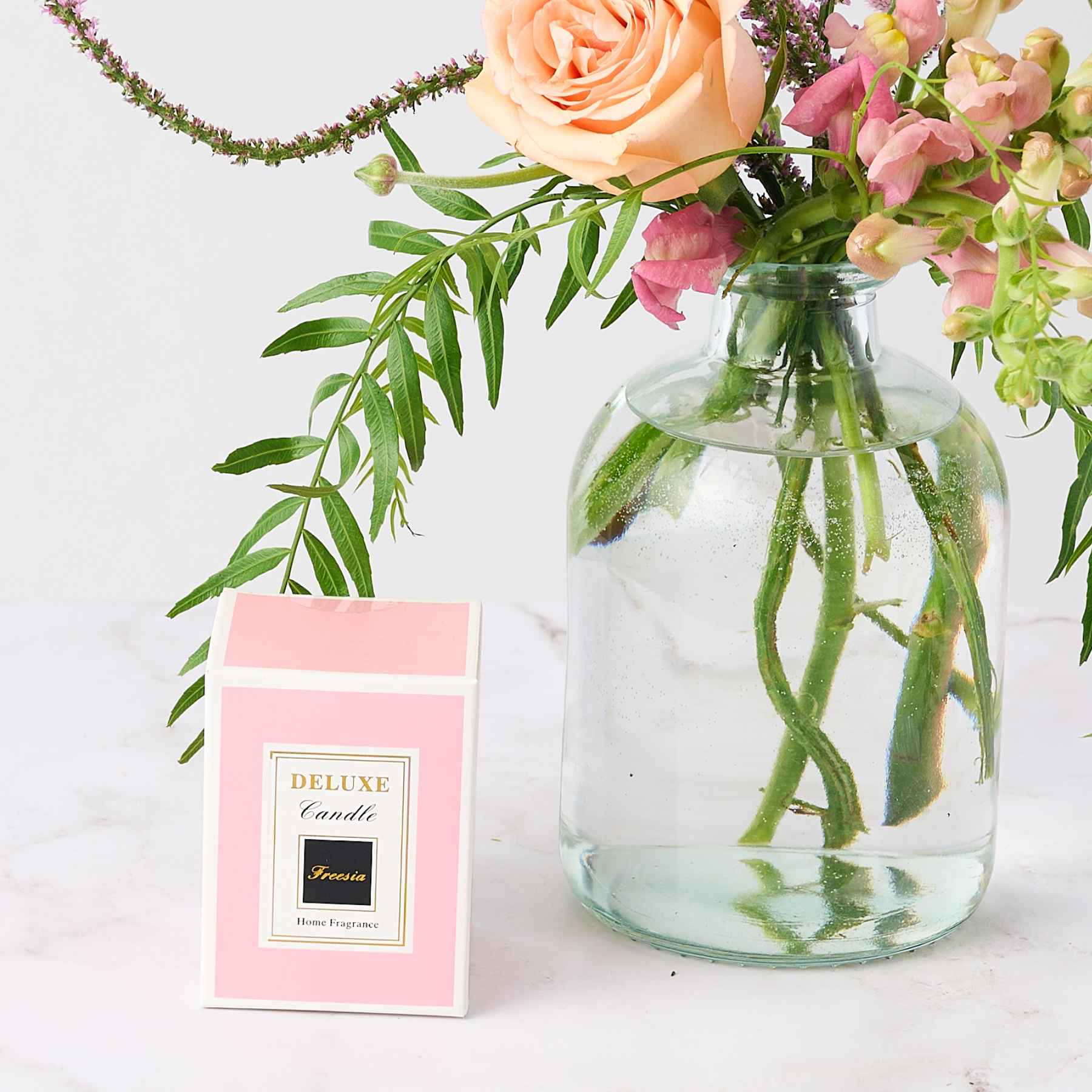 Close-up of a pink deluxe candle box next to a small vase with peach roses and pink flowers, representing the 'A Moment of Calm Pamper Gift Set'.