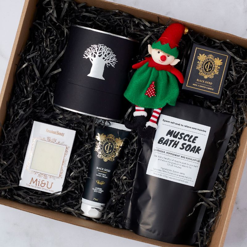 Gift box with bath essentials, including muscle bath soak, hand cream, artisan soap, candle, and a festive gnome plush.