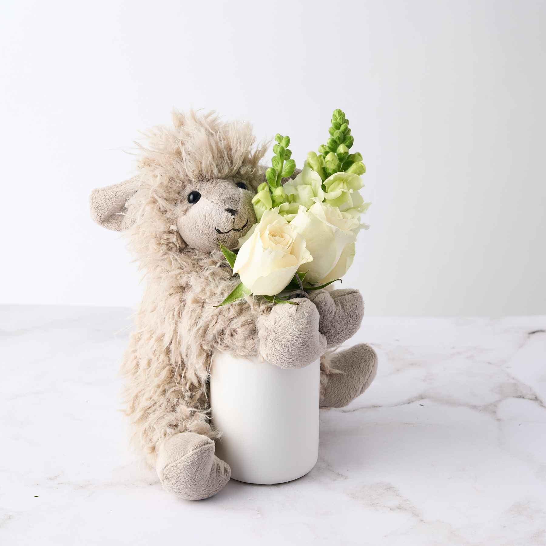 A beige plush toy holding a small vase with white roses and greenery, part of the 'Baby Girl Gift' arrangement.