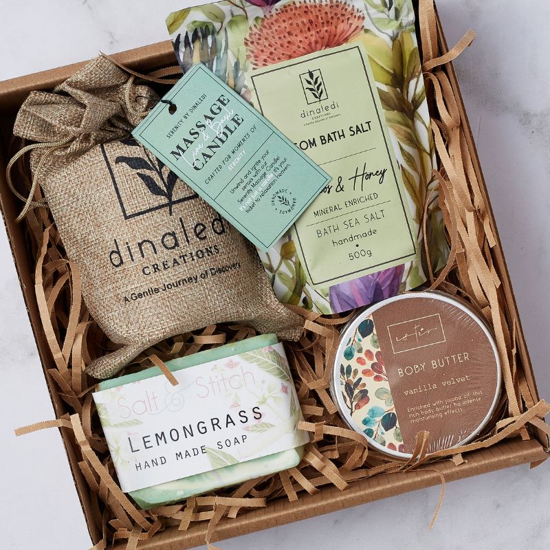 Close-up of the Blissful Indulgences gift box containing a Dinaledi massage candle, rooibos & honey bath salt, Salt & Stitch lemongrass handmade soap, and vanilla velvet body butter, arranged with shredded paper filler