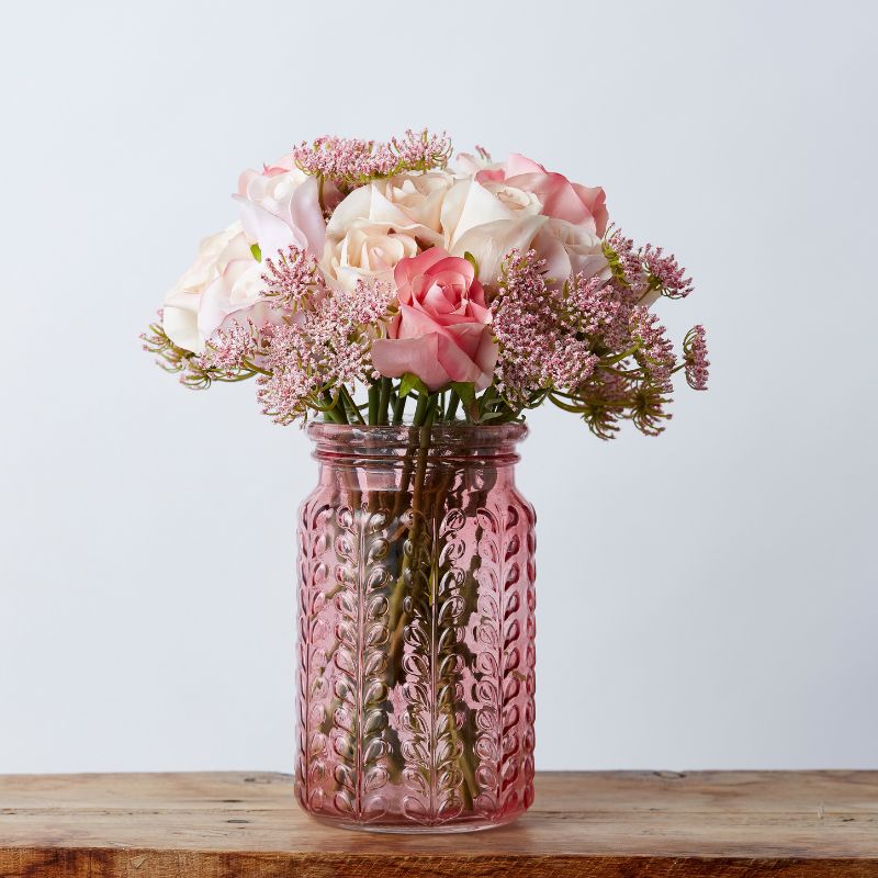 Artificial blush and cream rose bouquet in a textured pink glass vase, placed on a wooden surface.