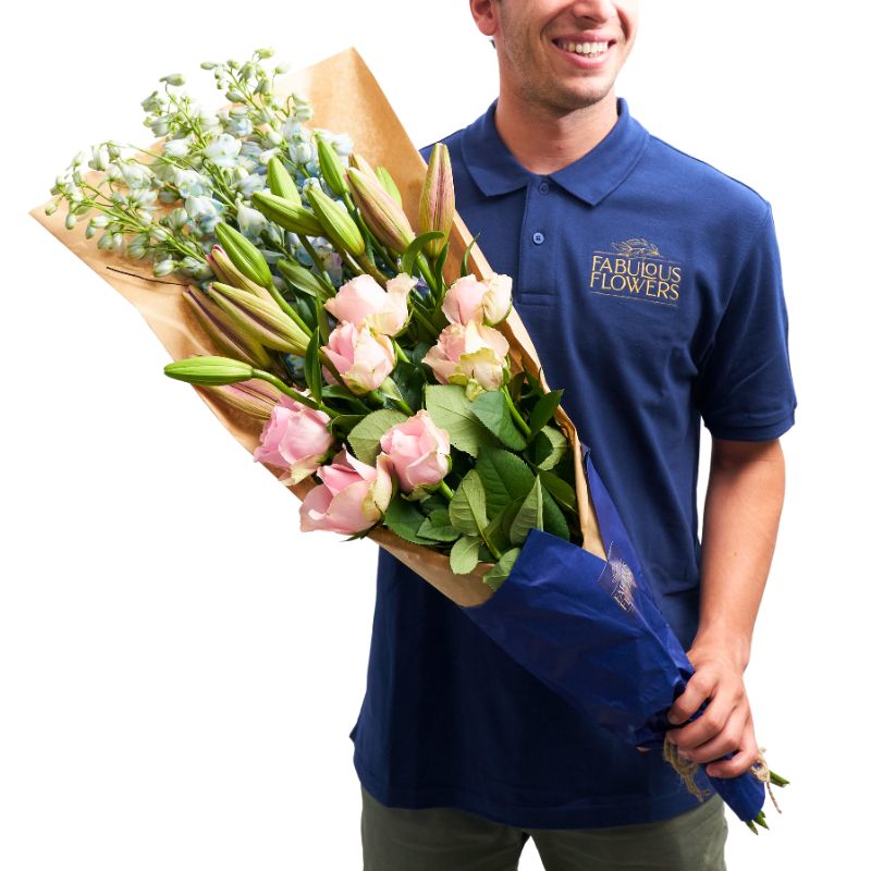 Blushing Beauty Bouquet from Fabulous Flowers & Gifts, featuring pink roses, lilies, and blue delphiniums wrapped in kraft paper.