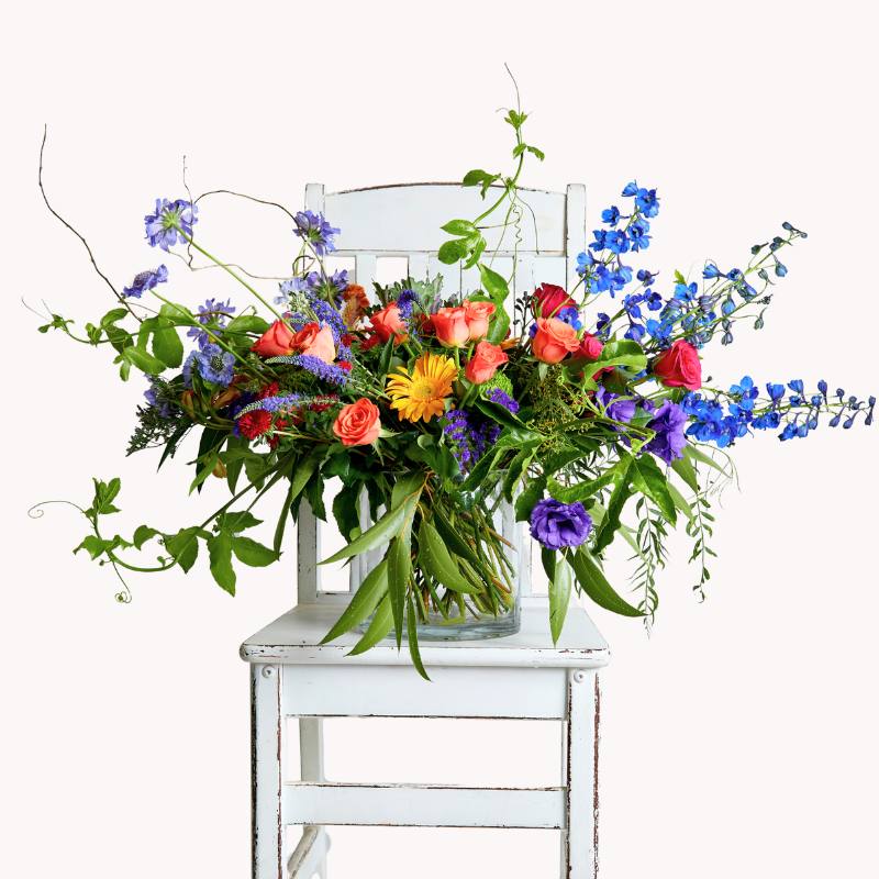 Vibrant flower arrangement on white chair, featuring roses, delphiniums, and lush greenery.