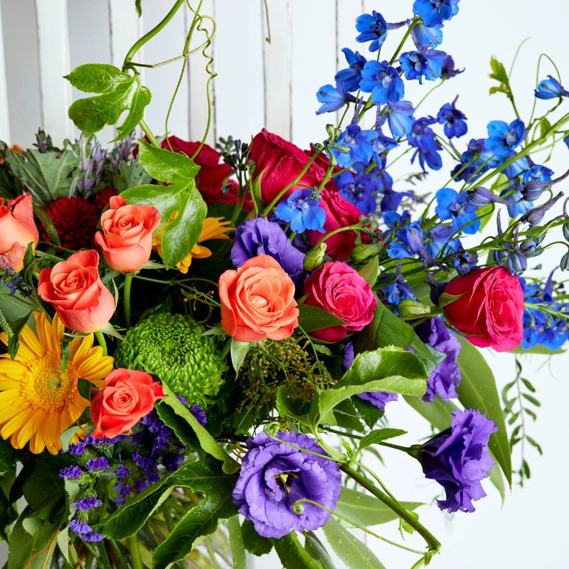 Close-up of colourful flower arrangement with roses, delphiniums, and a variety of green leaves.