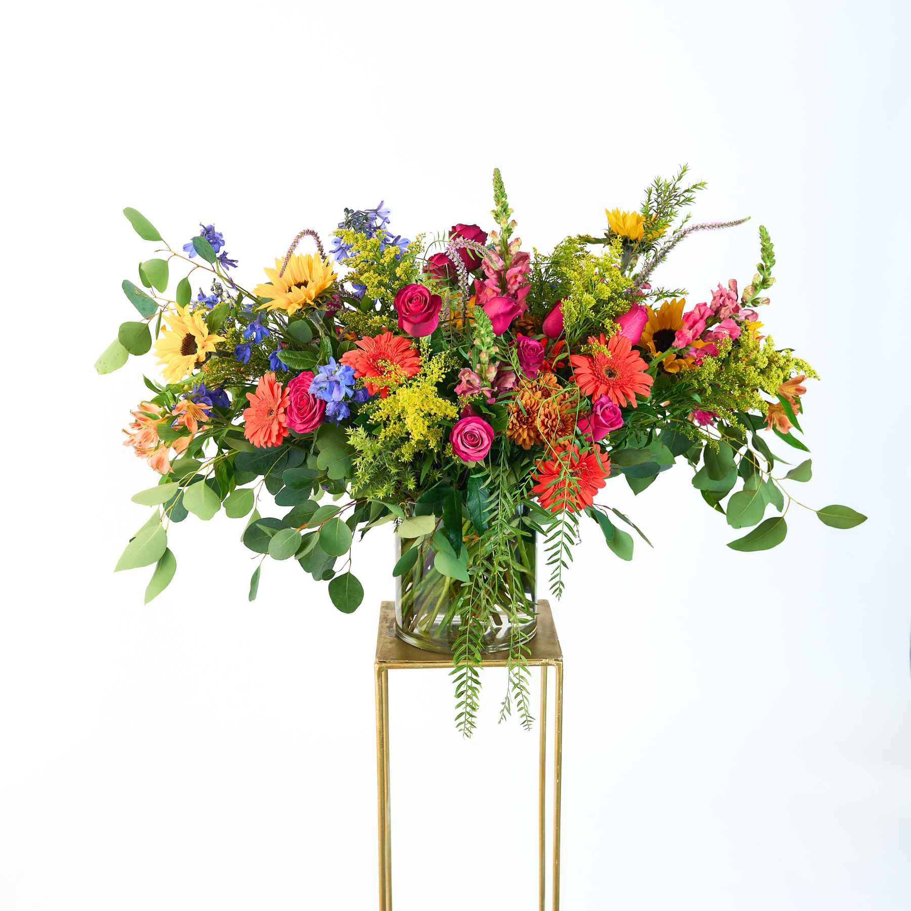 A vibrant and modern flower arrangement in a glass vase, featuring a bold mix of colourful flowers like roses, gerberas, sunflowers, and lush greenery, creating a lively and cheerful display