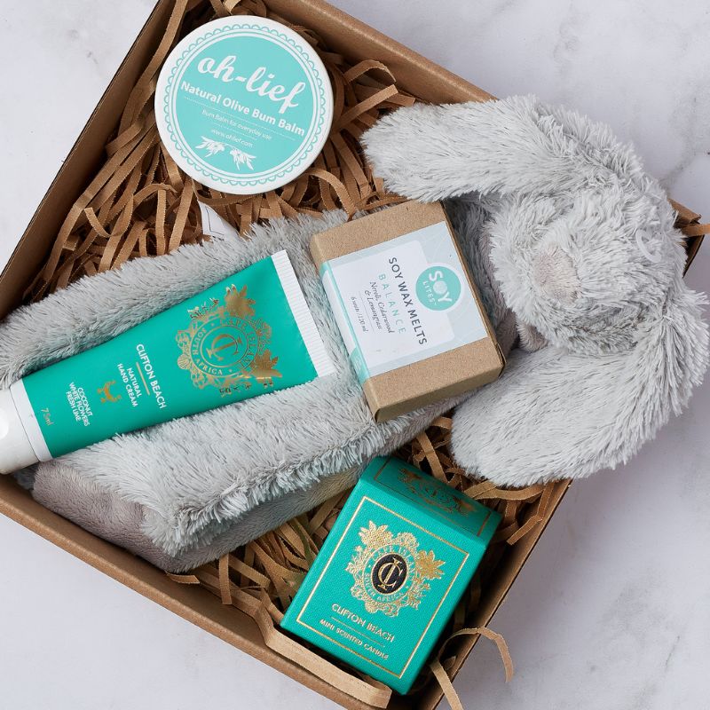 Close-up of gift box containing Oh-Lief bum balm, Clifton Beach hand lotion, soy wax melts, mini candle, and soft bunny toy.