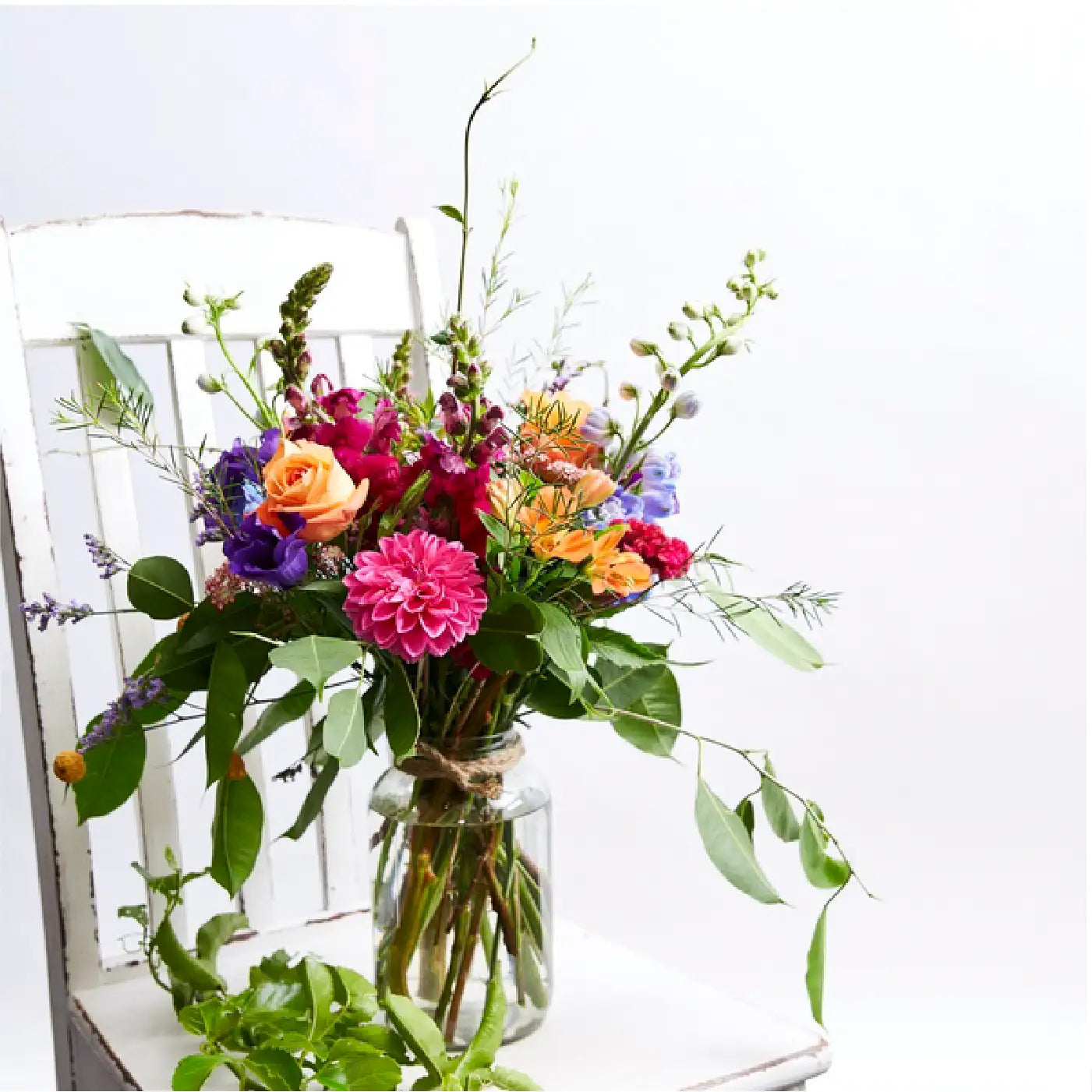 A vibrant bouquet of flowers in a glass jar, featuring orange roses, pink dahlias, purple lisianthus, and assorted greenery, placed on a white wooden chair. Business Gifting. Fabulous Flowers and Gifts.
