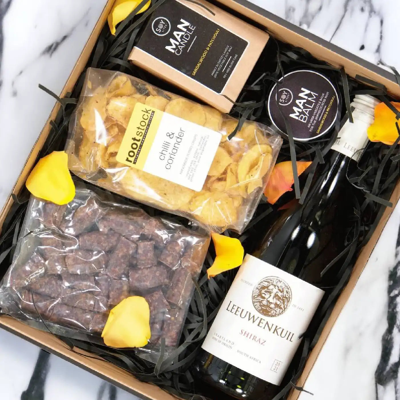 A luxurious gift box featuring a bottle of Leeuwenkuil Shiraz wine, chilli and coriander crisps, man balm, man candle, and a packet of gourmet nuts, arranged with yellow rose petals. Business Gifting. Fabulous Flowers and Gifts.