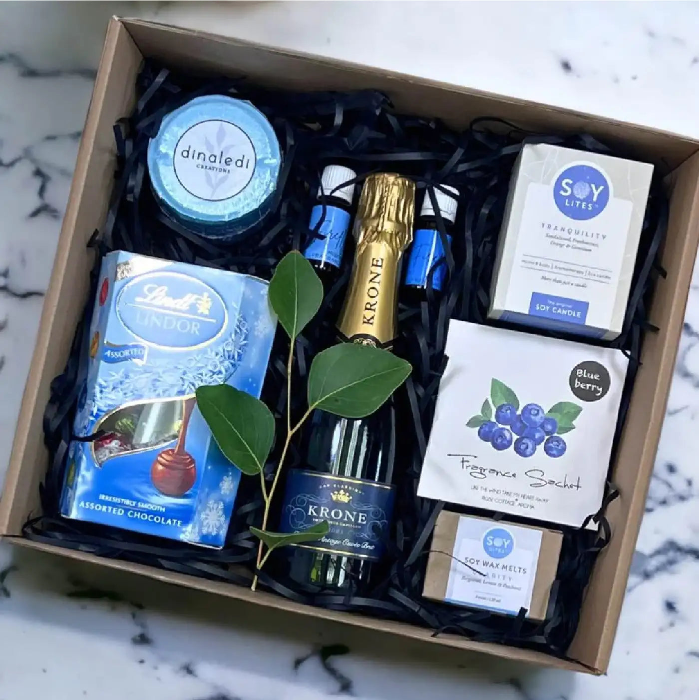 Elegant gift box featuring Lindt Lindor chocolates, Krone sparkling wine, soy candles, fragrance sachets, and wax melts, neatly arranged with a sprig of greenery on a marble background. Perfect for business gifting. Fabulous Flowers and Gifts.