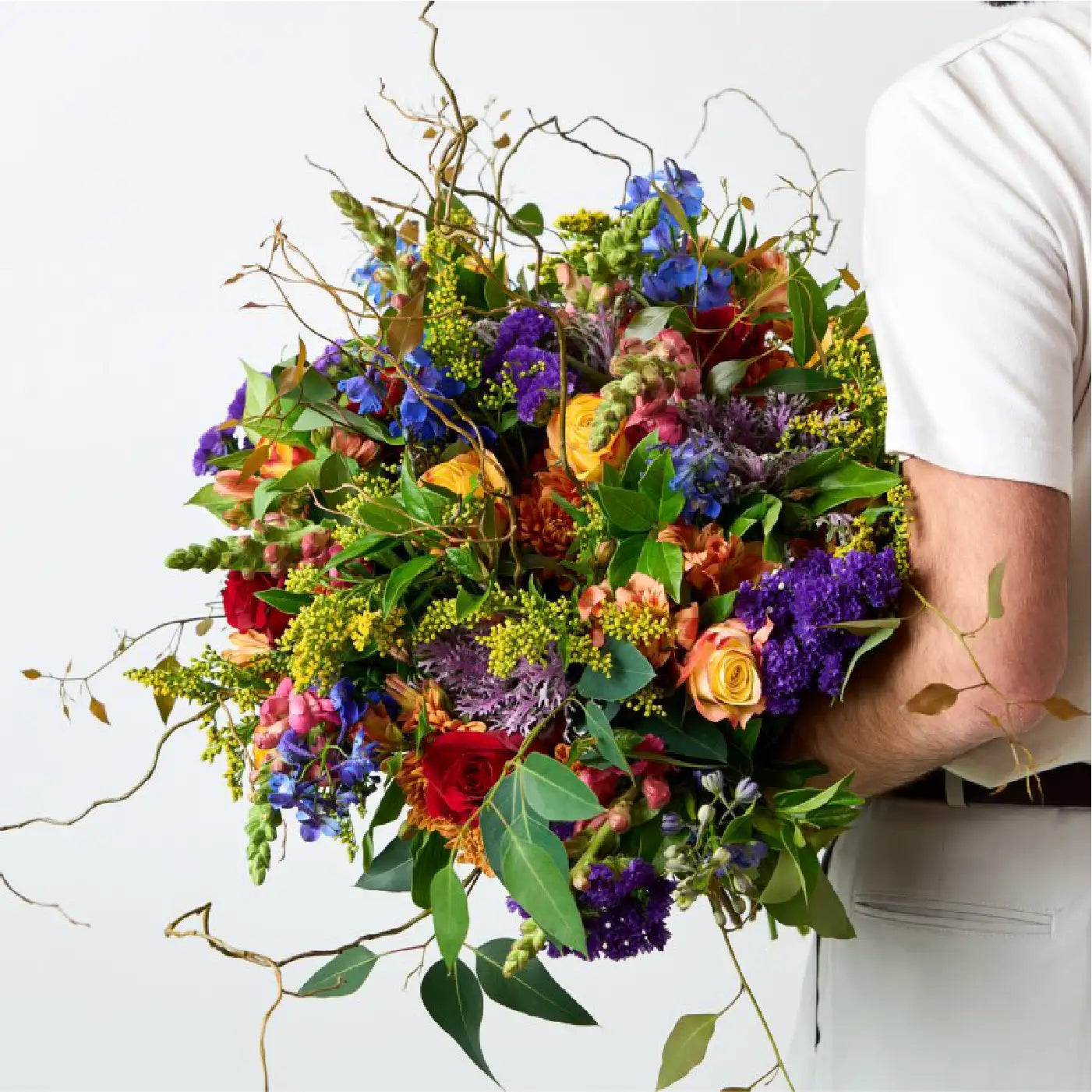 Person holding a large, vibrant bouquet of mixed flowers including roses, delphiniums, and greenery against a white background. Business Gifting. Fabulous Flowers and Gifts.