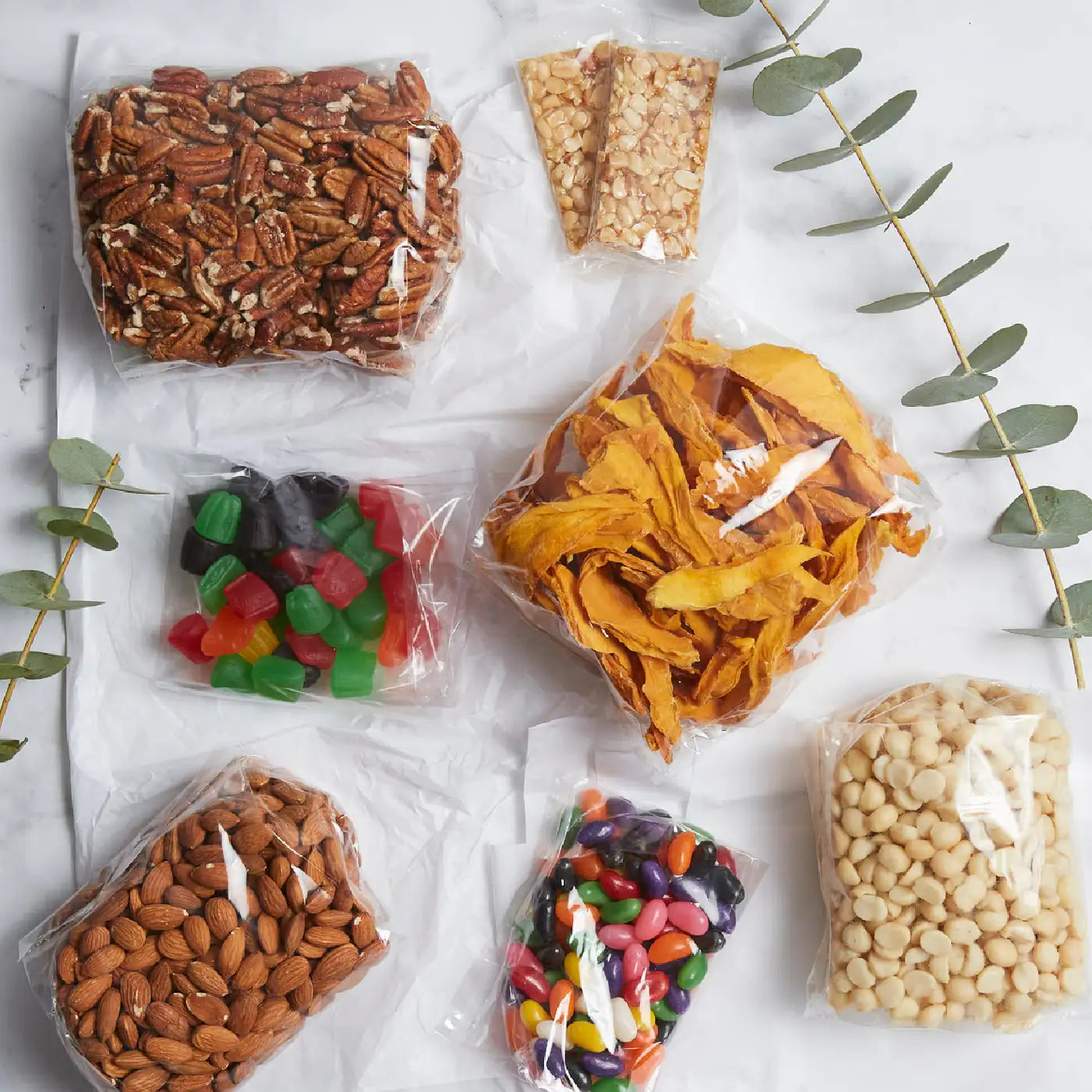 Assorted snack packs including nuts, dried mango, and sweets arranged on a white marble surface with eucalyptus leaves for a business gifting set. Business Gifting. Fabulous Flowers and Gifts.