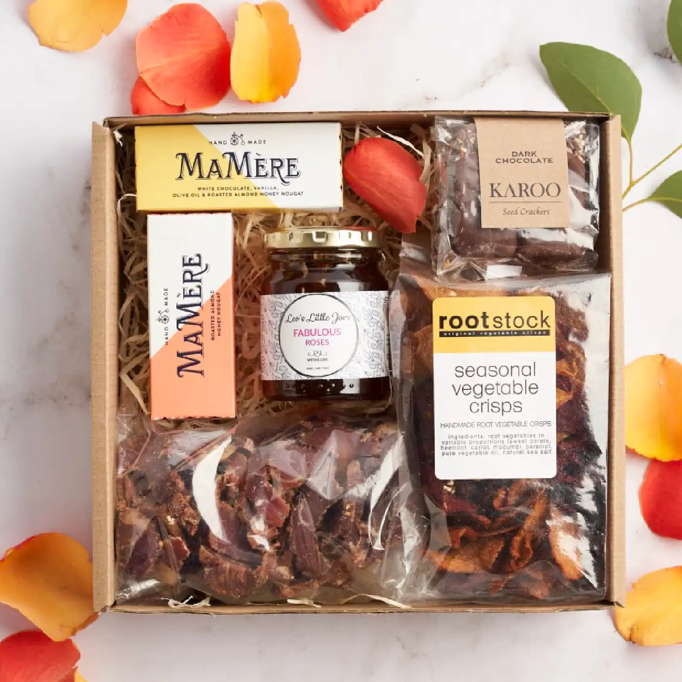 A curated business gifting box featuring gourmet items including MaMère nougat, seasonal vegetable crisps, dark chocolate seed crackers, a jar of rose jam, and biltong, elegantly arranged with decorative rose petals. Fabulous Flowers and Gifts.