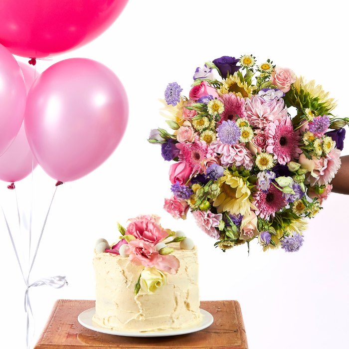 Close-up of a buttercream cake topped with pink roses and a lush floral bouquet held beside pink party balloons.