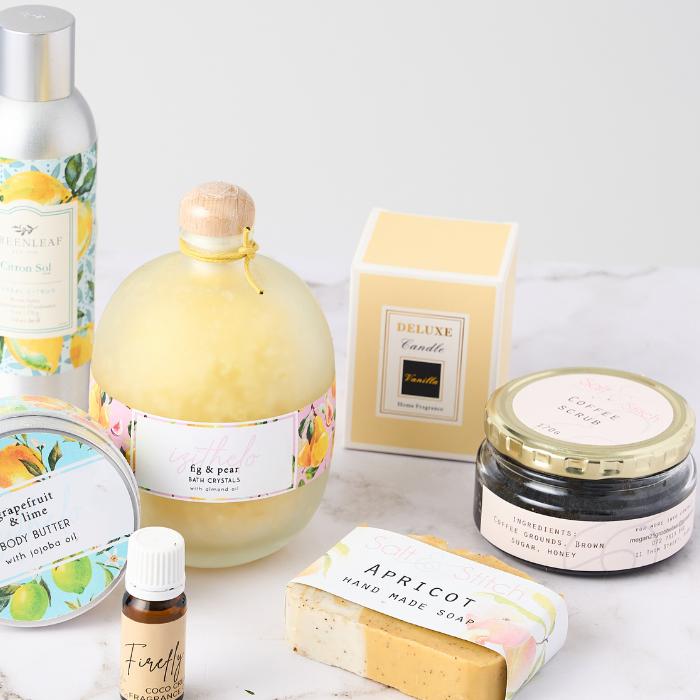  A collection of bath and body care products displayed on a white surface. The set includes fig & pear bath crystals in a round bottle with a wooden stopper, a deluxe vanilla candle in a yellow box, a coffee scrub in a glass jar with a gold lid, an apricot handmade soap bar, grapefruit & lime body butter in a metal tin, a small bottle of Firefly coconut fragrance oil, and a citrus-scented lotion in an aluminum bottle. The packaging features floral and citrus designs, evoking a fresh, natural, and luxurious 