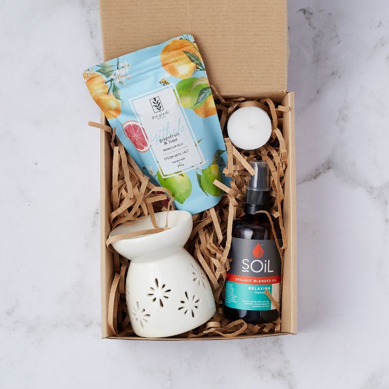 Gift box with a calming spa set, including grapefruit and lime bath salts, SOiL organic relaxing massage oil, ceramic oil burner, and tea light candle, arranged in shredded paper packaging.