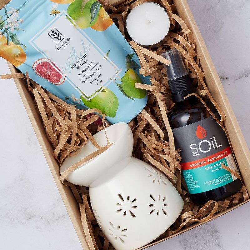 lose-up of a spa gift box with grapefruit and lime bath salts, SOiL organic relaxing massage oil, ceramic oil burner, and tea light candle, in shredded paper packaging.