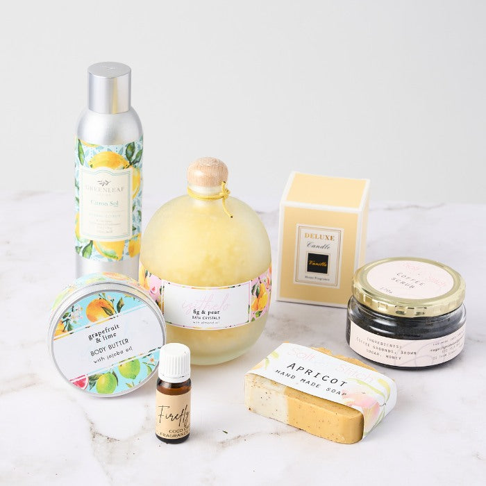 A selection of luxury bath and body care products arranged on a white surface. The products include a grapefruit & lime body butter in a tin, a fig & pear bath crystal bottle with a wooden top, a coffee scrub in a jar, an apricot handmade soap, a citrus-scented lotion in a tall aluminum bottle, a Firefly fragrance oil, and a yellow box containing a deluxe candle. The packaging features colorful floral and citrus designs, evoking a fresh, natural, and vibrant feel.