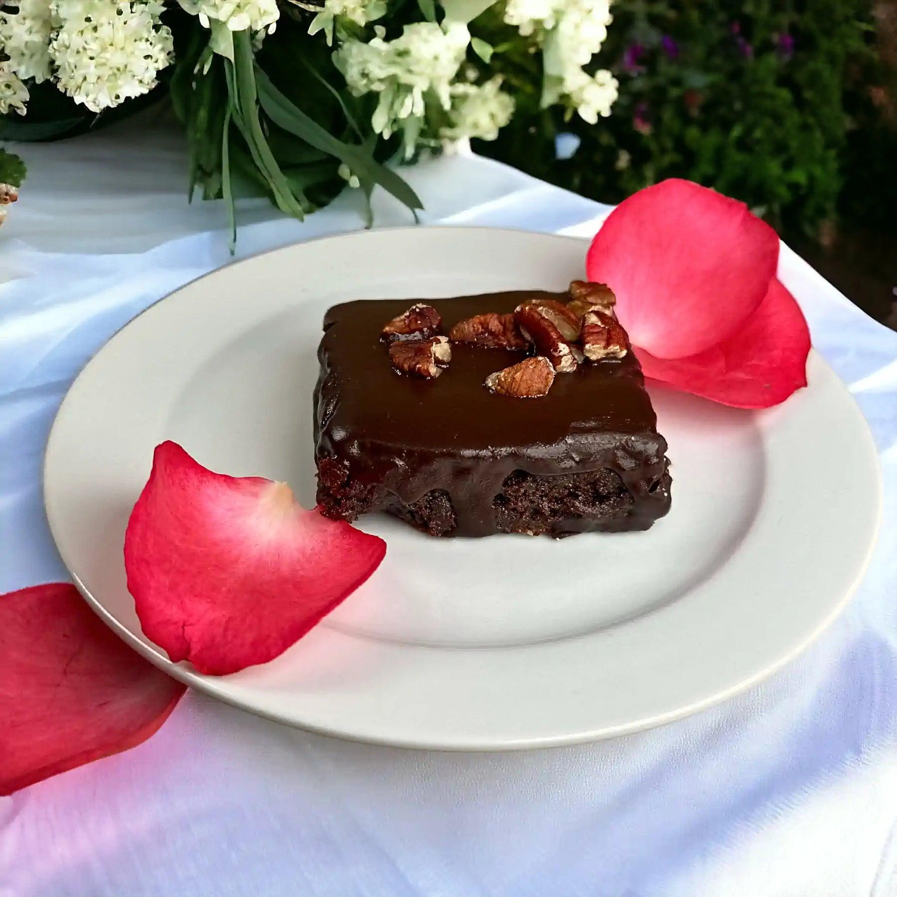 Rich gluten-free chocolate brownie topped with pecans and adorned with red rose petals on a white plate. Fabulous Flowers and Gifts.