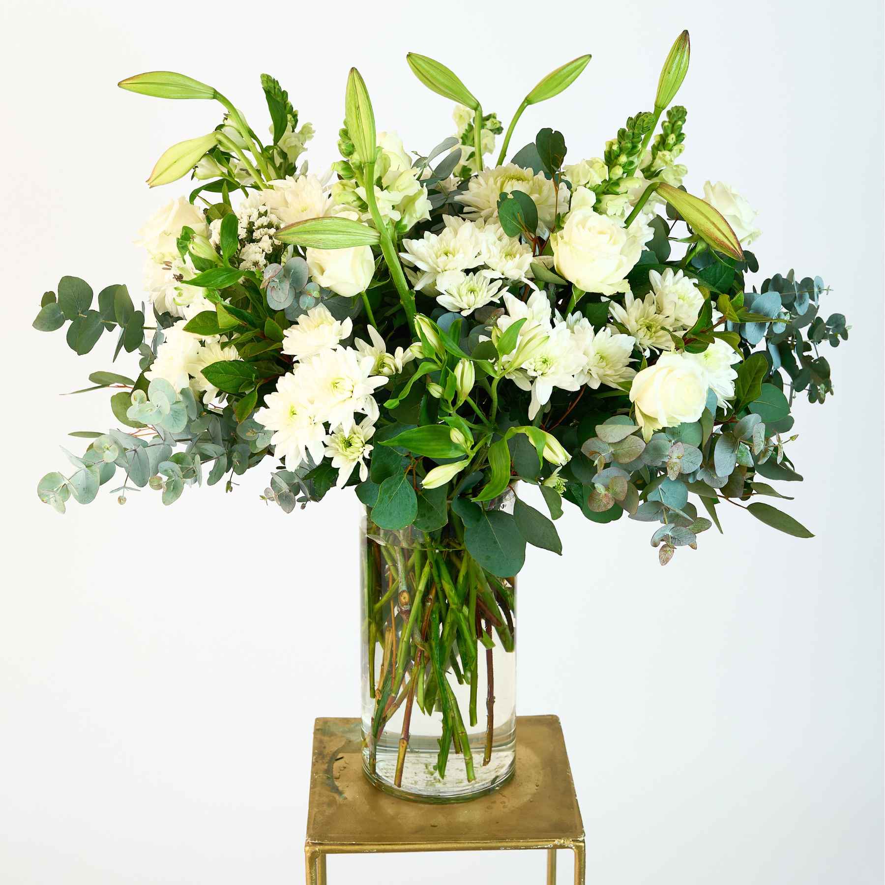 Elegant white flower arrangement in a glass vase, featuring roses, lilies, and lush green foliage, displayed on a minimalistic stand.