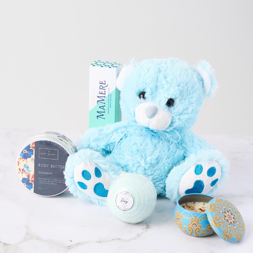 Cozy Cuddles Gift Set featuring a plush blue teddy bear, bergamot body butter, bath bomb, candle, and Mamère nougat.