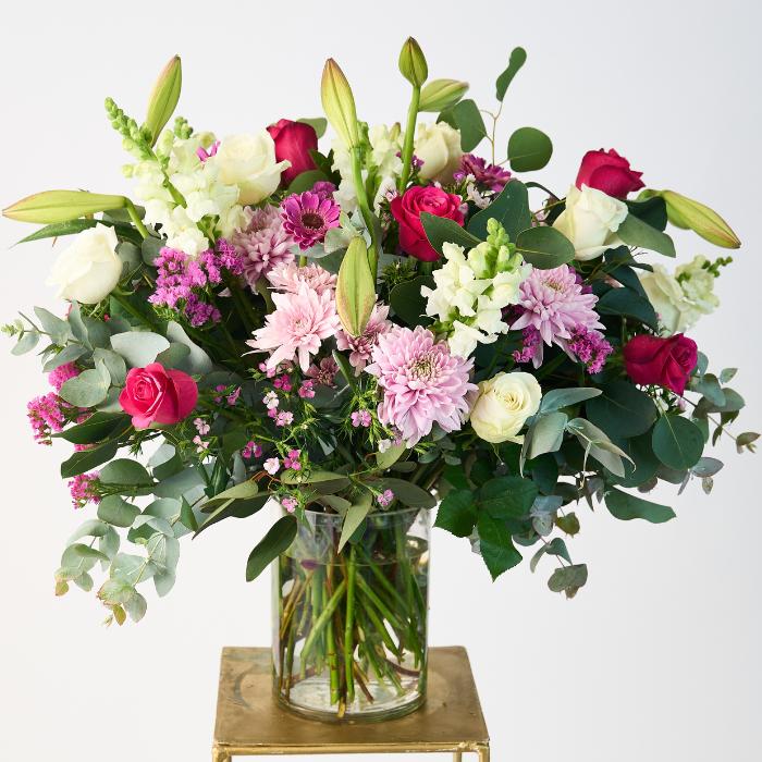 Creative Elegance Arrangement featuring a lush bouquet of vibrant pink and white roses, gerberas, lilies, and wild greenery in a clear glass vase, expertly arranged to create a captivating and elegant display.