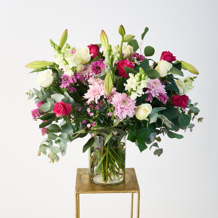 Creative Elegance Arrangement featuring a lush bouquet of vibrant pink and white roses, gerberas, lilies, and wild greenery in a clear glass vase, expertly arranged to create a captivating and elegant display.