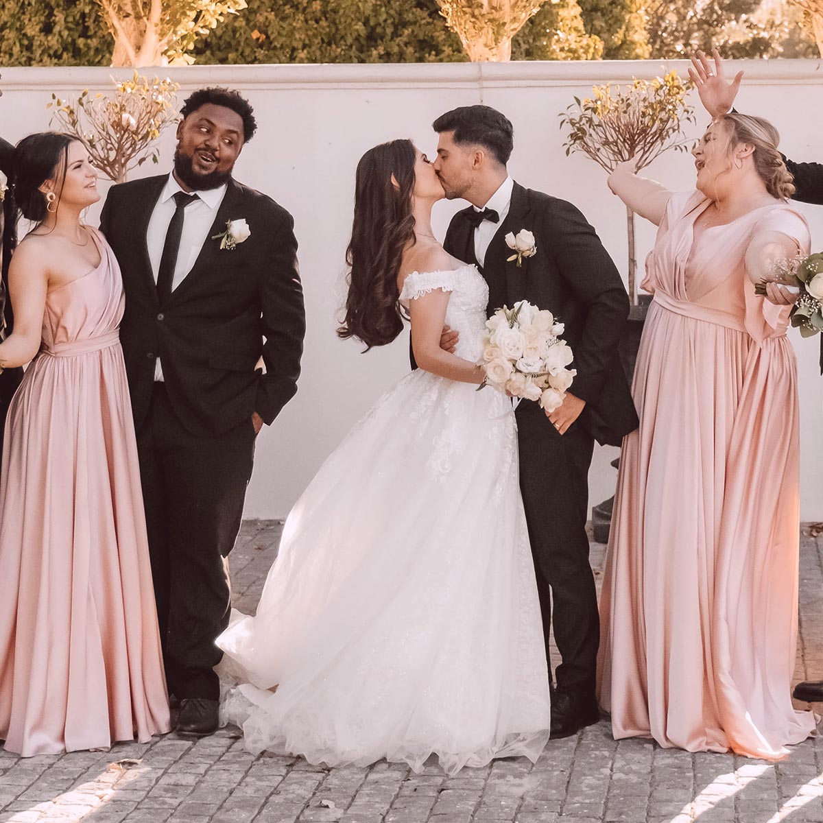 Bride and groom, Emilio and Carmen, share a kiss surrounded by their joyful bridesmaids and groomsman in elegant attire. Weddings. Fabulous Flowers and Gifts.