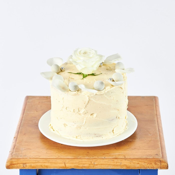 Elegant Rose Celebration Cake featuring creamy vanilla frosting, a white rose on top, and decorative silver pearls, presented on a wooden table for a special occasion.