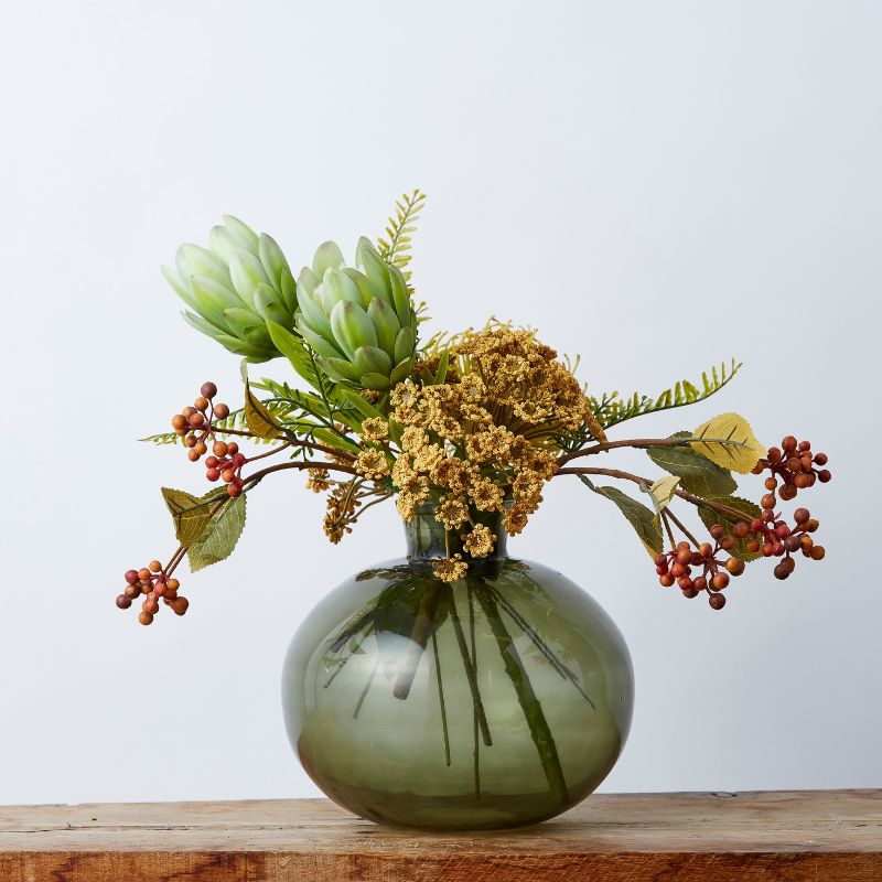 Elegant silk flower arrangement with green proteas, golden clusters, red berries, and lush greenery in a sleek green vase.