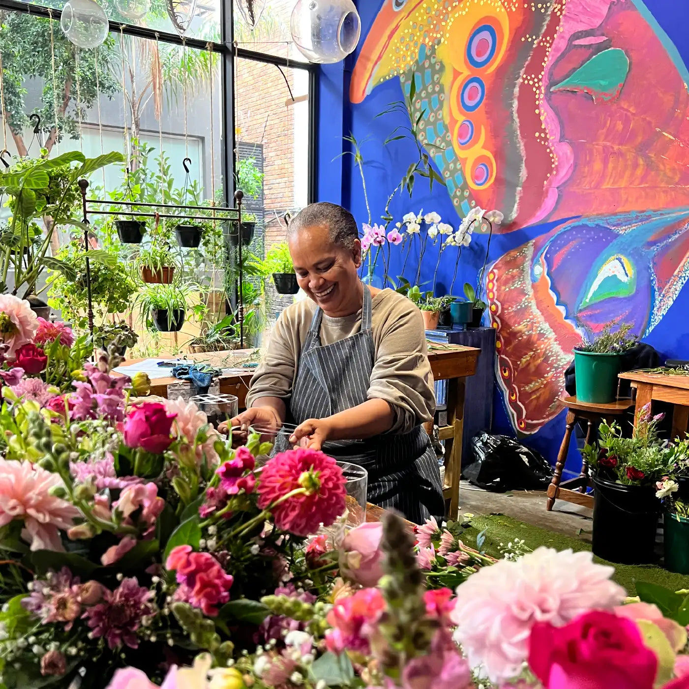 Smiling florist in a vibrant studio arranging a bouquet of colourful flowers with a butterfly mural in the background. Frequently Asked Questions. Fabulous Flowers and Gifts.
