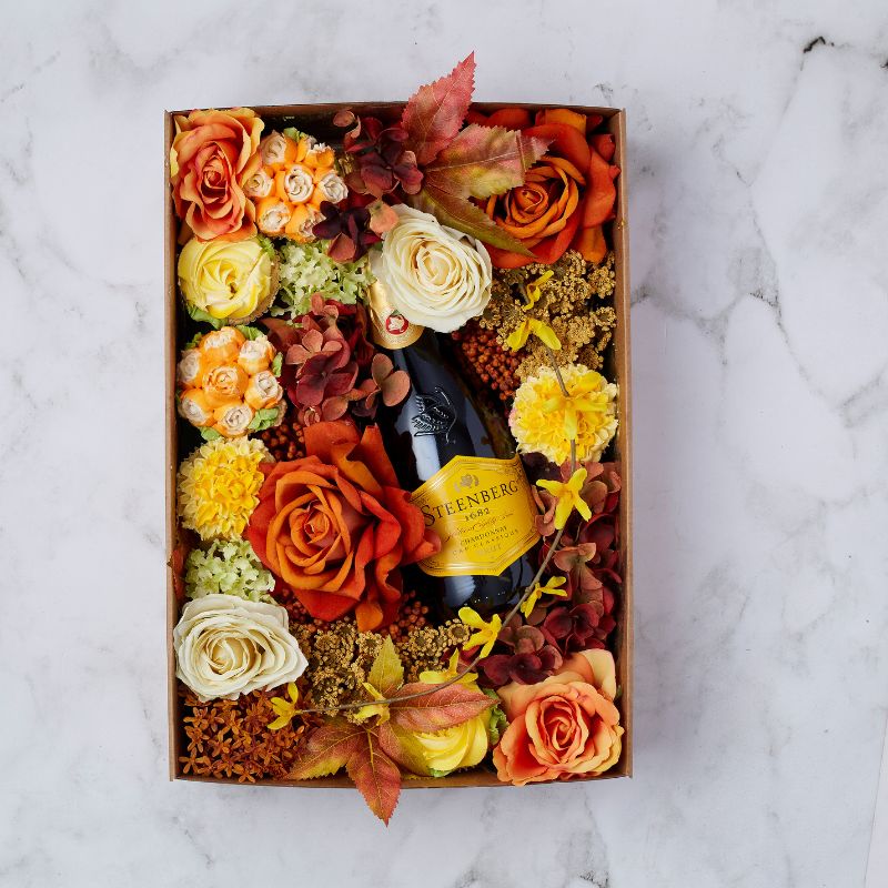 Autumn-themed gift box with Steenberg Chardonnay Brut surrounded by vibrant orange and yellow flowers.