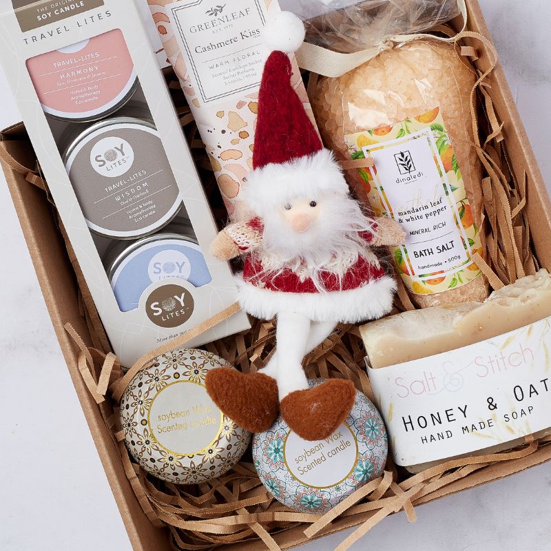 Christmas gift box featuring a festive Santa plush toy, Soy Lites travel candle set, Greenleaf Cashmere Kiss sachet, Dinaledi Mandarin Leaf & White Pepper bath salts, Salt & Stitch Honey & Oat handmade soap, and two decorative soybean wax scented candles.