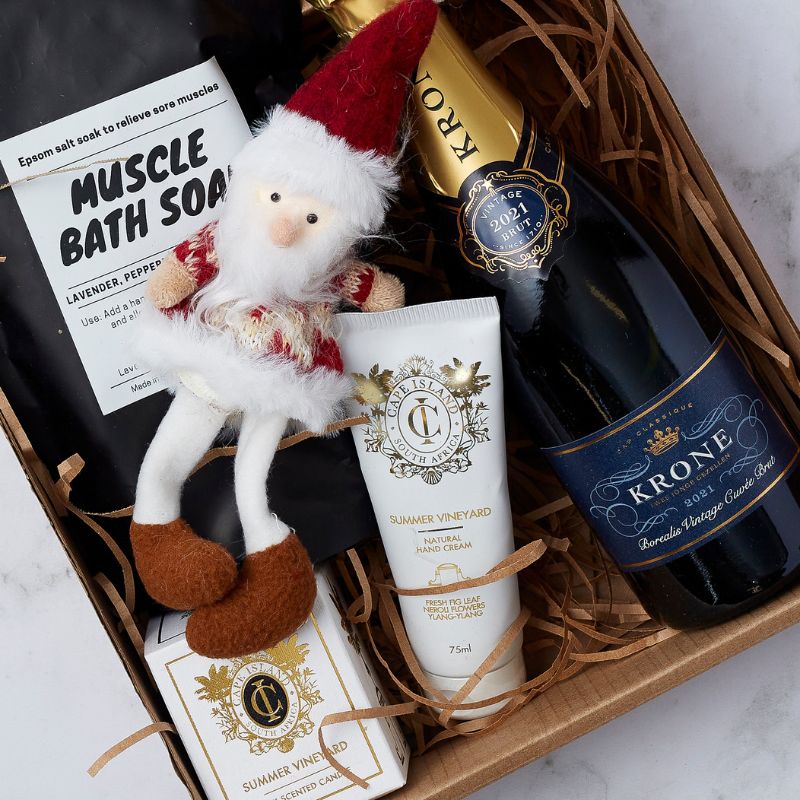 Festive gift hamper featuring Krone sparkling wine, muscle bath soak, Cape Island hand cream and candle, and Santa plush decoration.