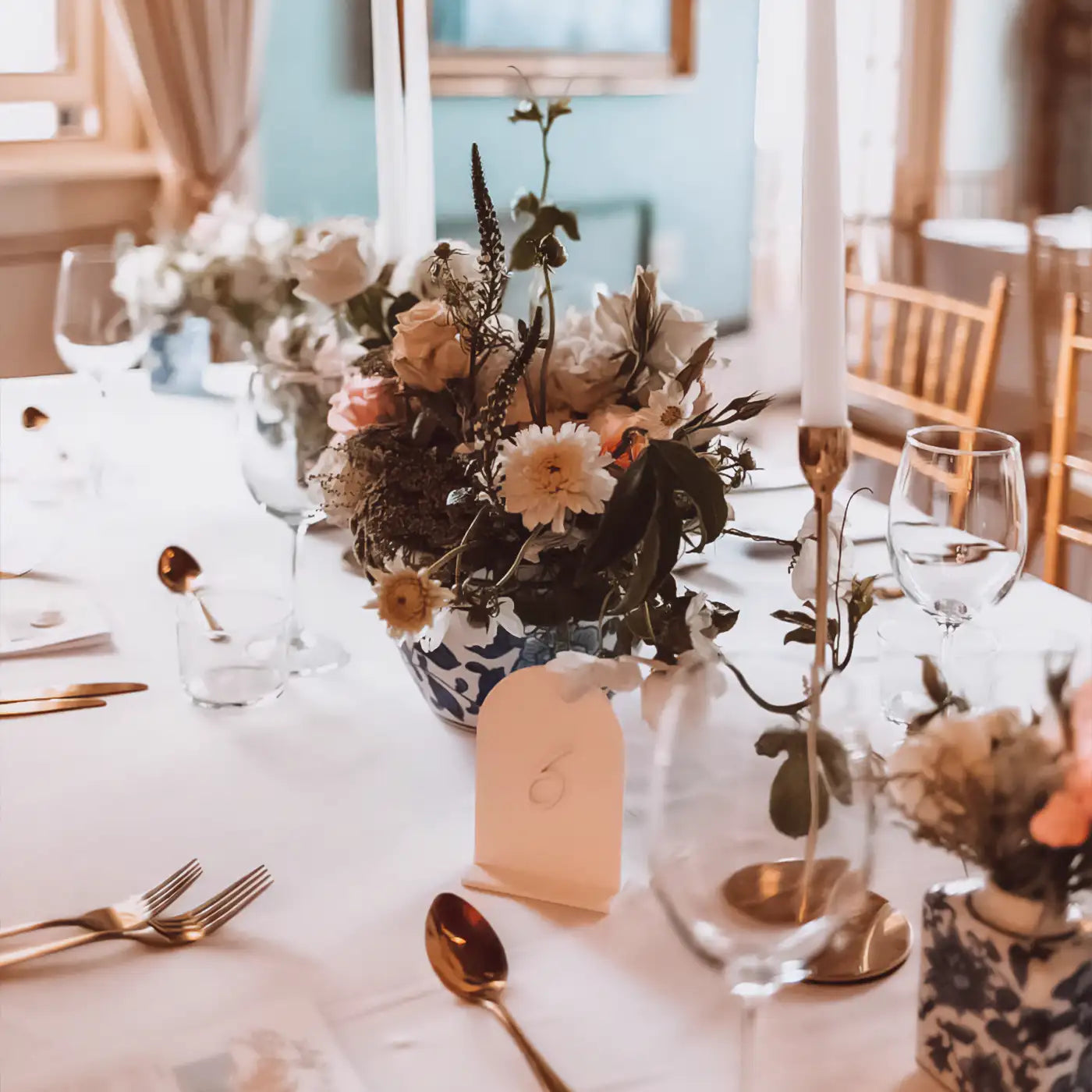 Close-up of a beautifully set table at an elegant event with white linens, gold cutlery, glassware, and blue and white floral centrepieces in vases, complemented by tall candlesticks. Fabulous Flowers and Gifts.
