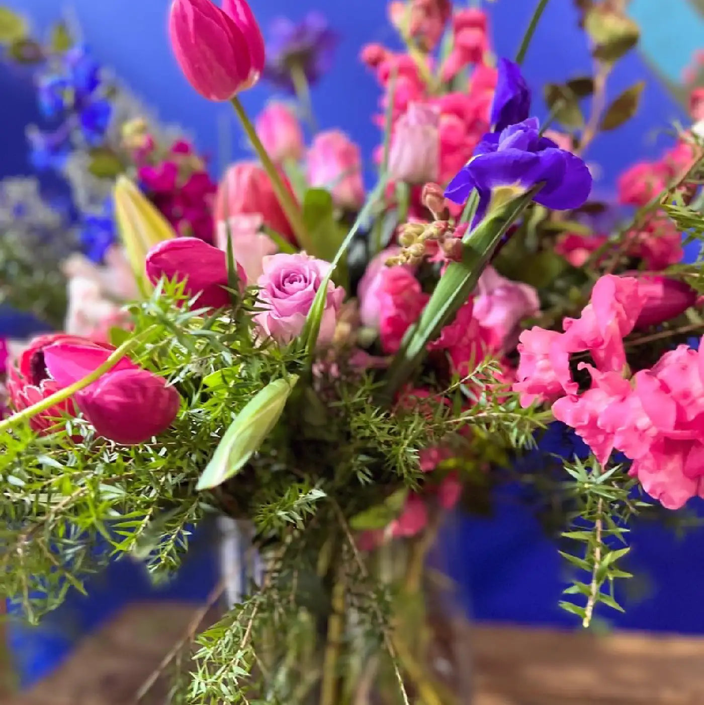Vibrant floral arrangement featuring pink roses, purple irises, and greenery against a blue background. Transform your space with Fabulous Flowers and Gifts. Flower Installations.