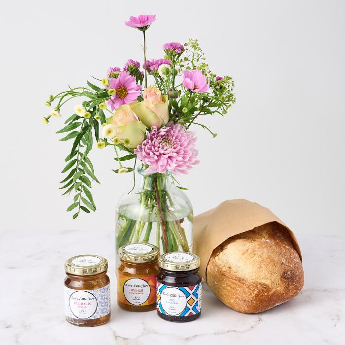 A glass vase filled with a vibrant bouquet of pink, purple, and white flowers, placed beside a loaf of freshly baked bread wrapped in brown paper, and three jars of artisanal jam labeled 'Fabulous Fables,' 'Orange,' and 'Fig.' Set on a light-colored surface, this arrangement exudes a warm and inviting atmosphere, perfect for a cozy gift.