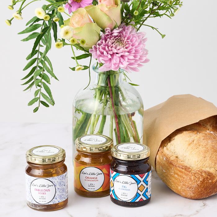 A glass vase filled with a beautiful arrangement of pink, purple, and peach flowers, displayed next to a loaf of fresh bread wrapped in brown paper and three jars of artisanal jam. The jams, labeled 'Fabulous Roses,' 'Orange & Cardamom,' and 'Fig & Lavender,' are set on a light surface, evoking a charming and cozy gift package.