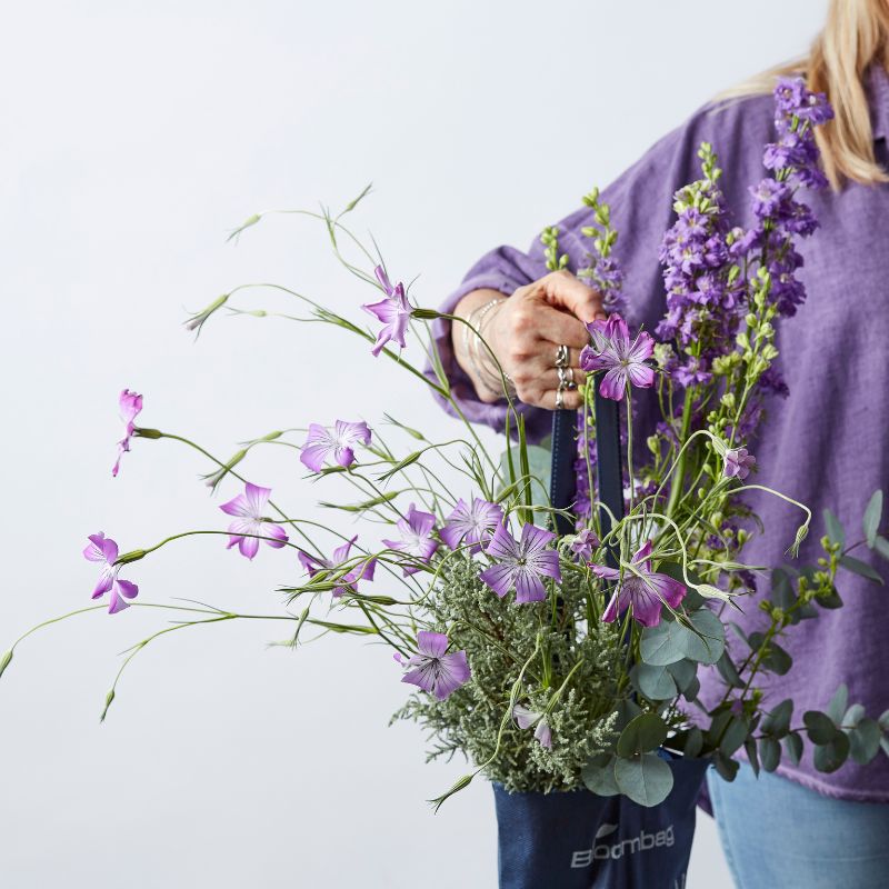 Close-up of a hand holding a bouquet of purple wildflowers and greenery in a blue reusable bag.