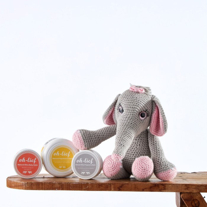 Gentle Giant Skincare Bundle featuring a handmade crochet elephant with grey and pink details, alongside natural olive pregnancy balm and baby balms, on a wooden surface.