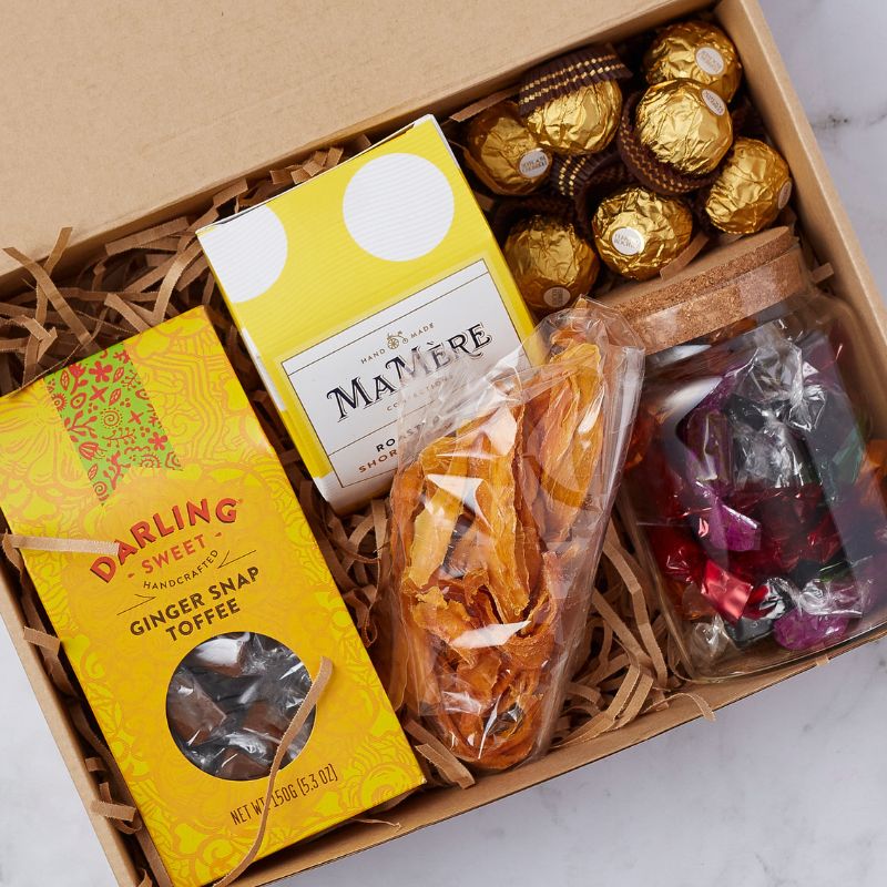 Close-up of an artisan treat box showcasing ginger snap toffee, Mamere shortbread, Ferrero Rocher chocolates, dried fruit, and a jar of colourful wrapped candies.