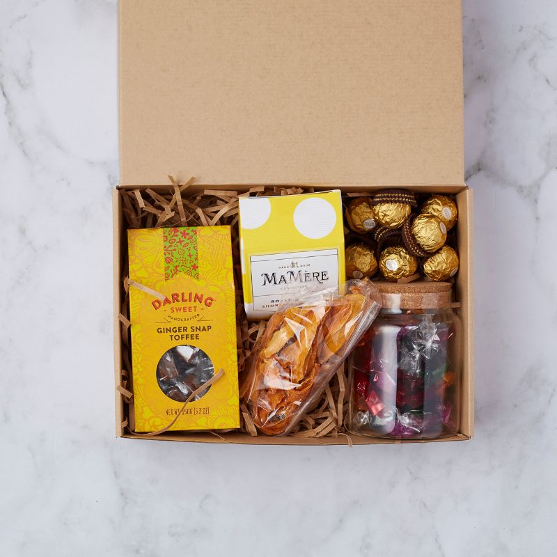 Top view of an artisan treat box featuring ginger snap toffee, Mamere shortbread, Ferrero Rocher chocolates, dried fruit, and a jar of colourful sweets.