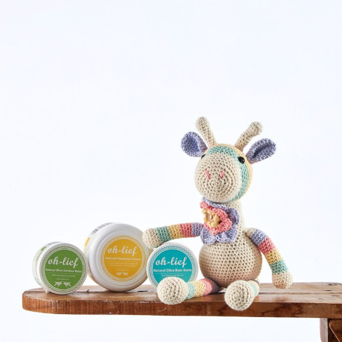 Handmade crochet giraffe with organic baby care balms including outdoor, aqueous, and olive bum balms on a wooden shelf – ideal for gentle newborn care.